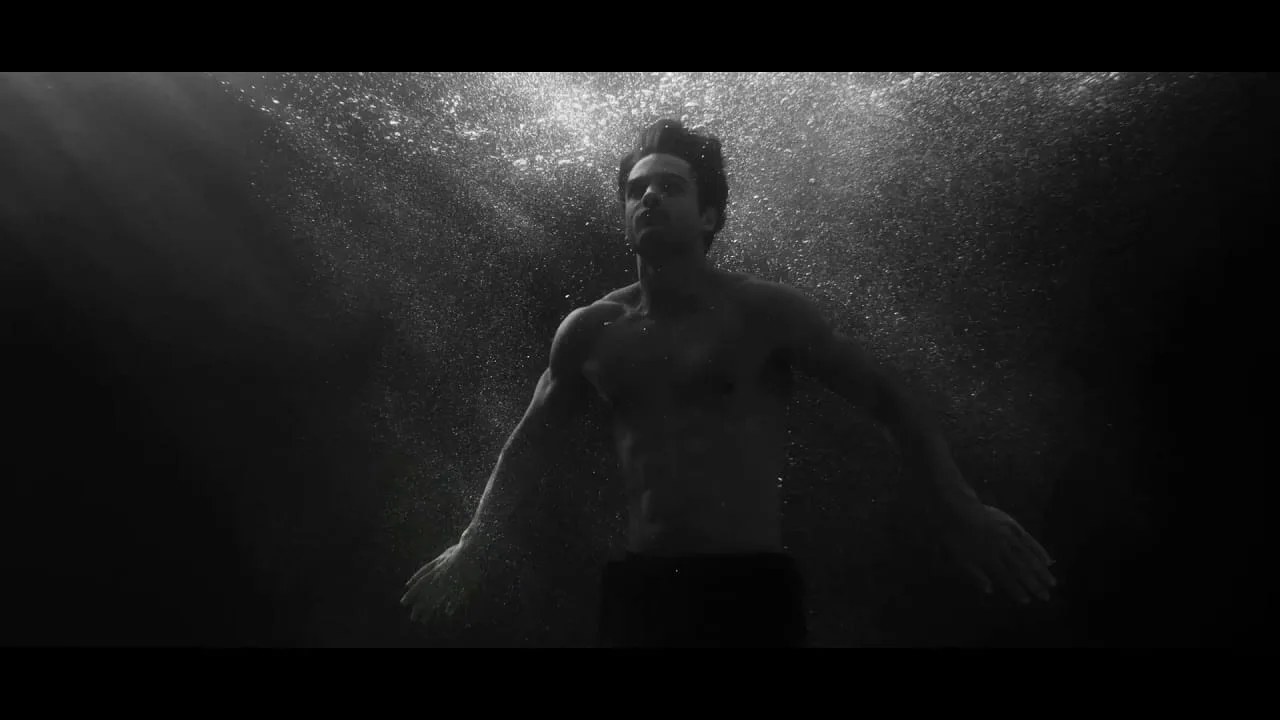 The swimmer suspended in black water just beneath the surface, arms spread, a galaxy of bubbles blooming above him where he broke through