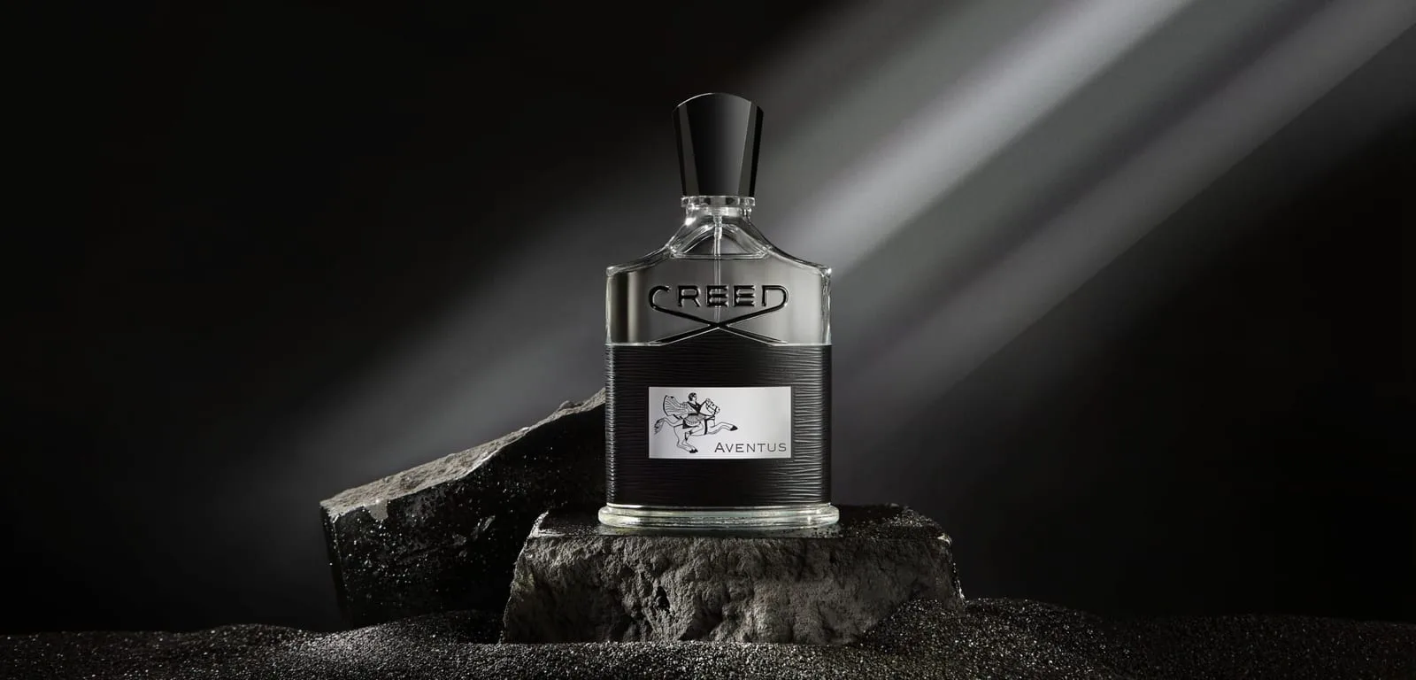 The Aventus flacon on a pedestal of raw black stone, shafts of pale light raking through the darkness behind it