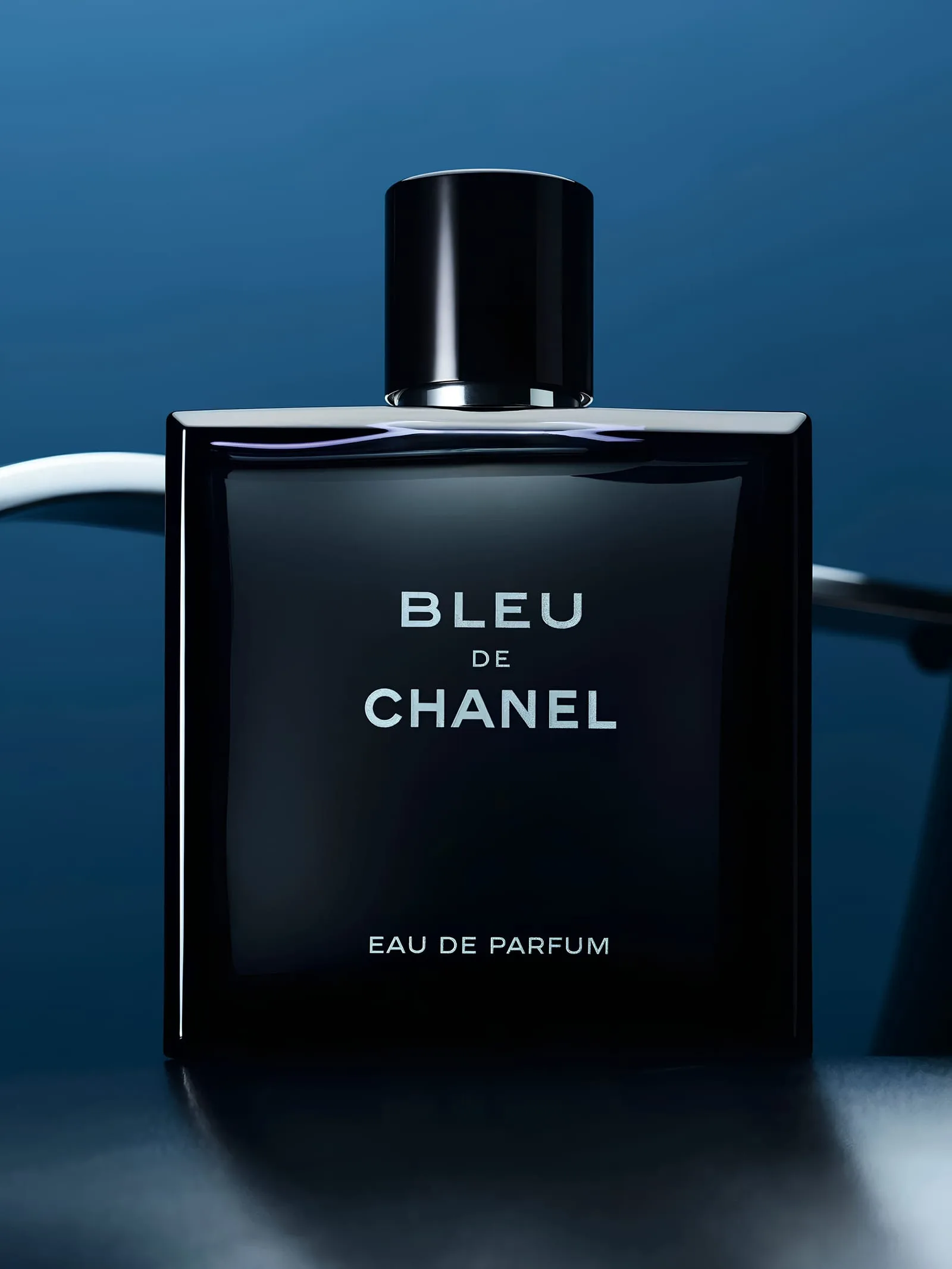The Eau de Parfum flacon stands alone against a slate-blue dusk, silver lettering catching the light, its reflection pooling on the dark surface below