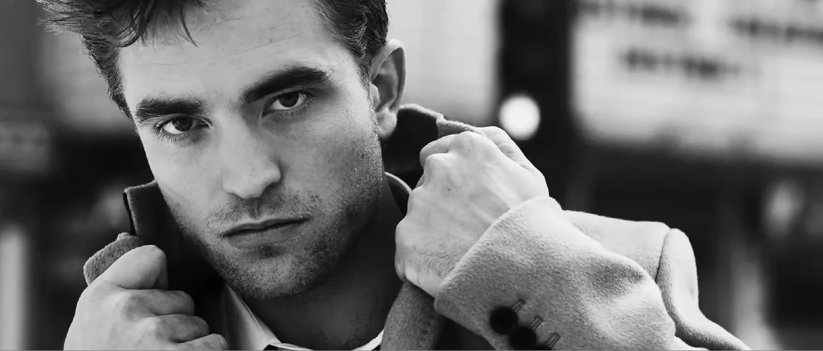 Robert Pattinson in black and white, collar of a camel overcoat gripped in both fists, a theatre marquee dissolving into grain behind him