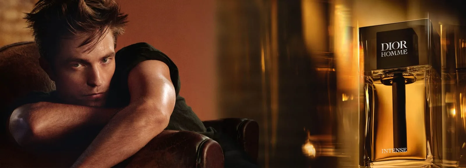 The Dior Homme Intense flacon stands hero-lit at right, burning amber against the smeared golden light of a darkened bar, Robert Pattinson watching from the leather shadows