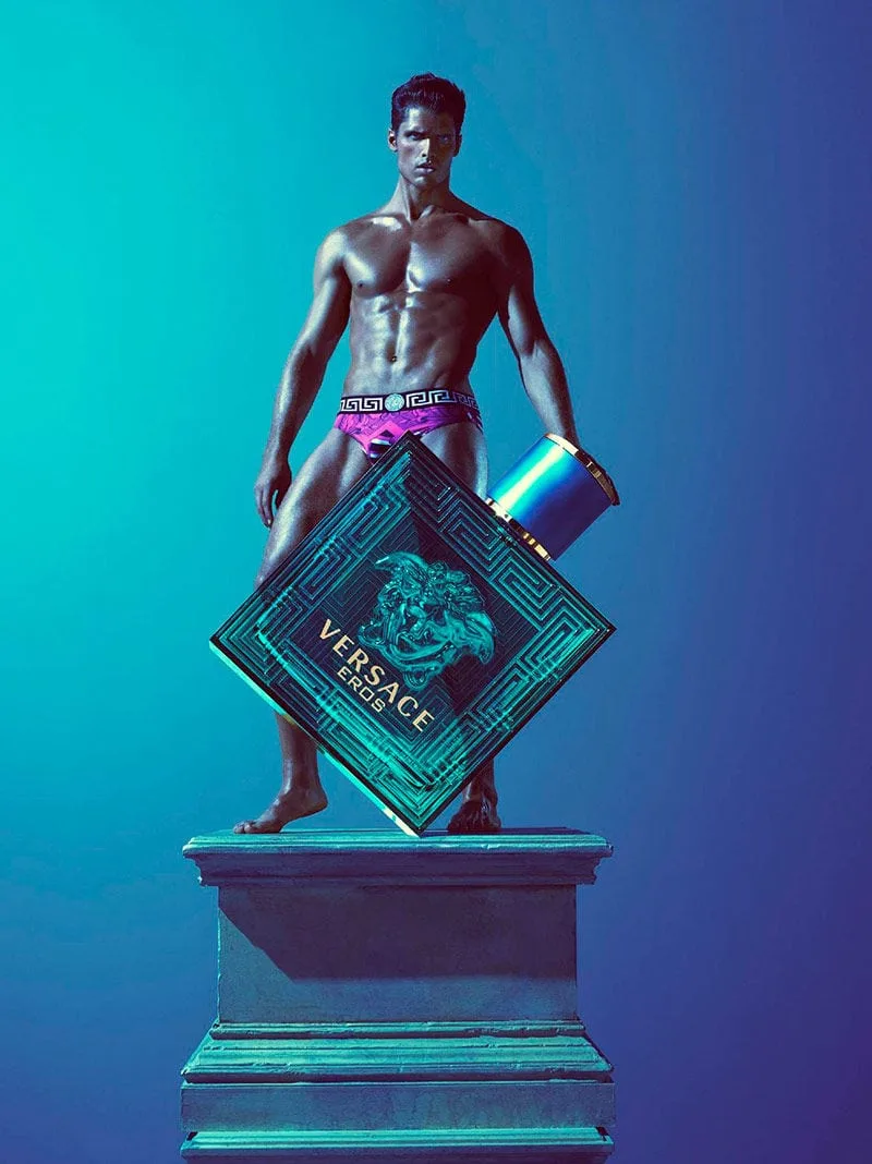 A living statue: the model stands barefoot atop a stone plinth, lit like patinated bronze in the campaign's chromatic blue, the giant bottle tipped at its base