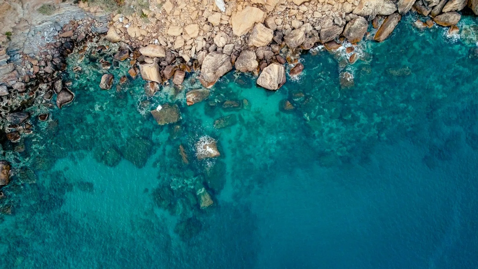 Aegean shallows seen from above at noon — turquoise water the exact colour of the Eros glass, breaking white over sun-bleached rock