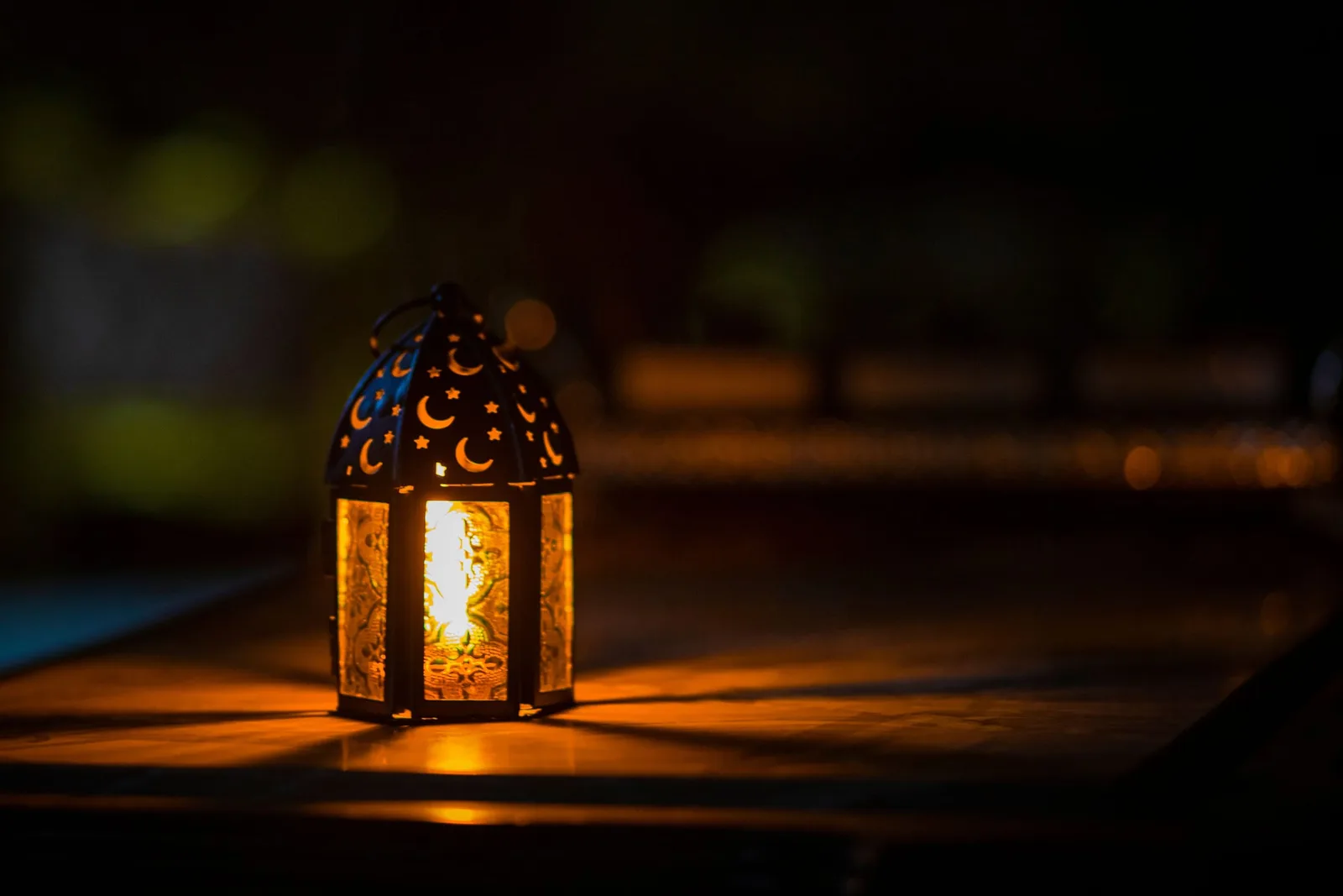 An Arabian lantern pierced with crescents and stars burns low on a dark table, spilling cinnamon-gold light across the wood while the night blurs to black around it