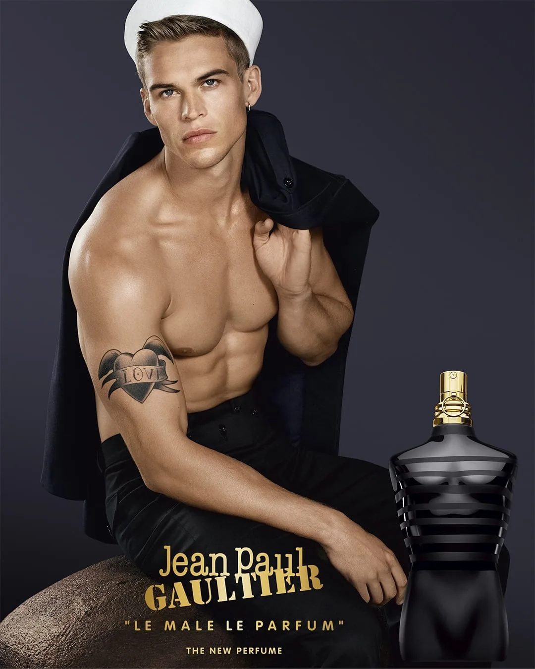The official poster: a sailor with white cap tilted back and a navy peacoat over bare shoulders sits in near-darkness beside the black-and-gold torso flacon
