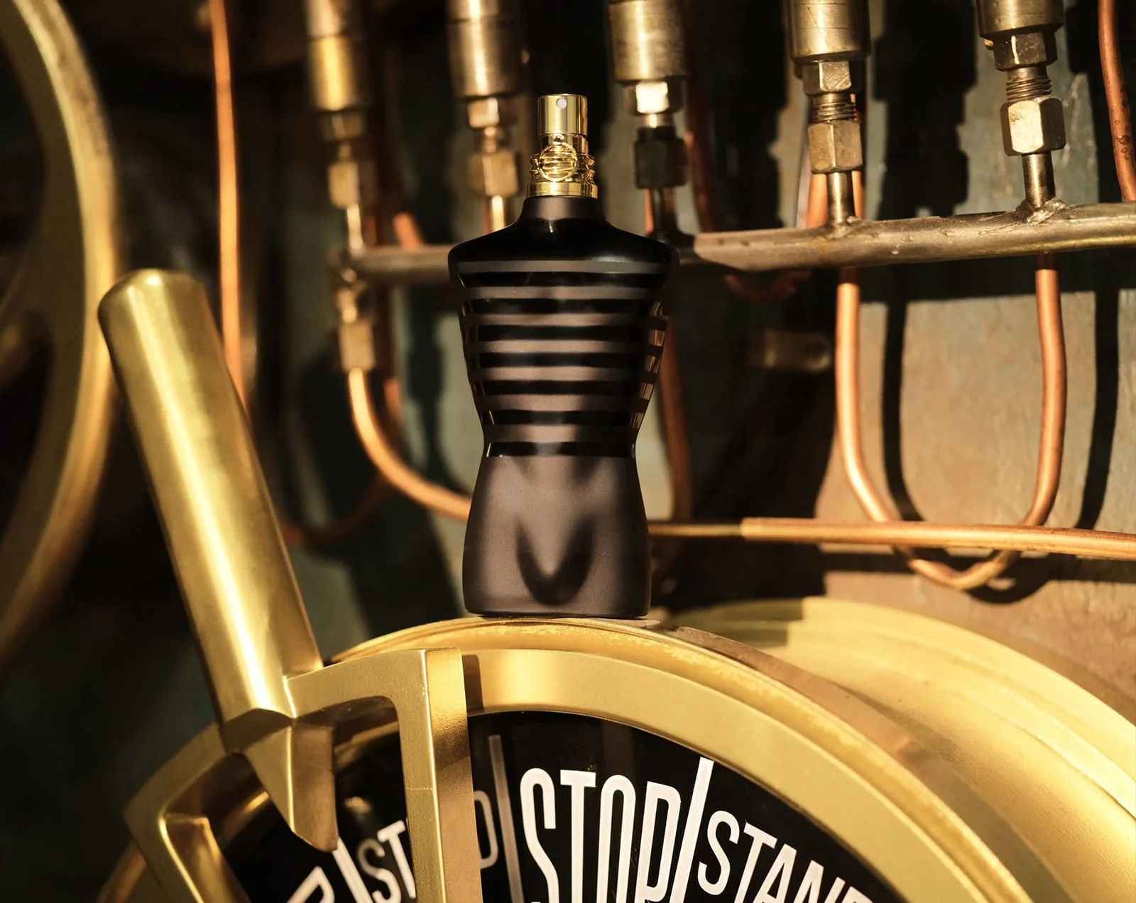 The matte-black striped torso, crowned in polished gold, stands at attention on the brass engine-order telegraph of a ship's engine room, amid copper pipework and lamplit valves