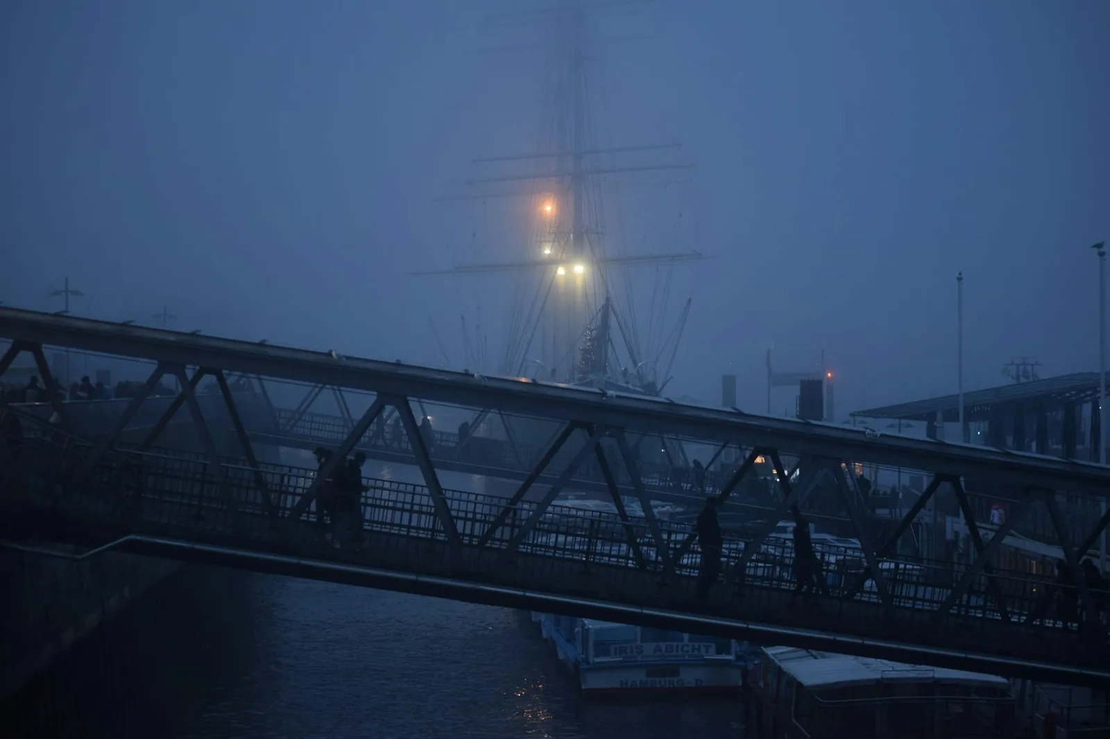 A tall ship's rigging dissolving into harbour fog after dark, two amber lamps burning warm against the blue-black night