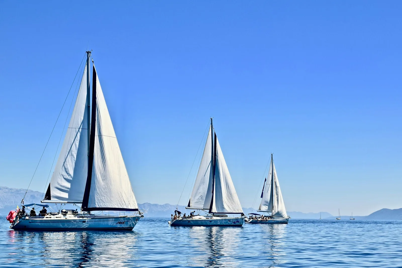 Three white sails leaning into a brilliant Mediterranean noon, the sea a deep lacquered blue and the far islands fading to haze