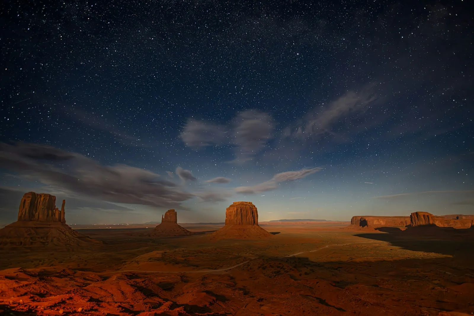 The buttes of Monument Valley standing sentinel under a sky thick with stars, moonlight tracing their edges while the desert floor holds a faint ember glow