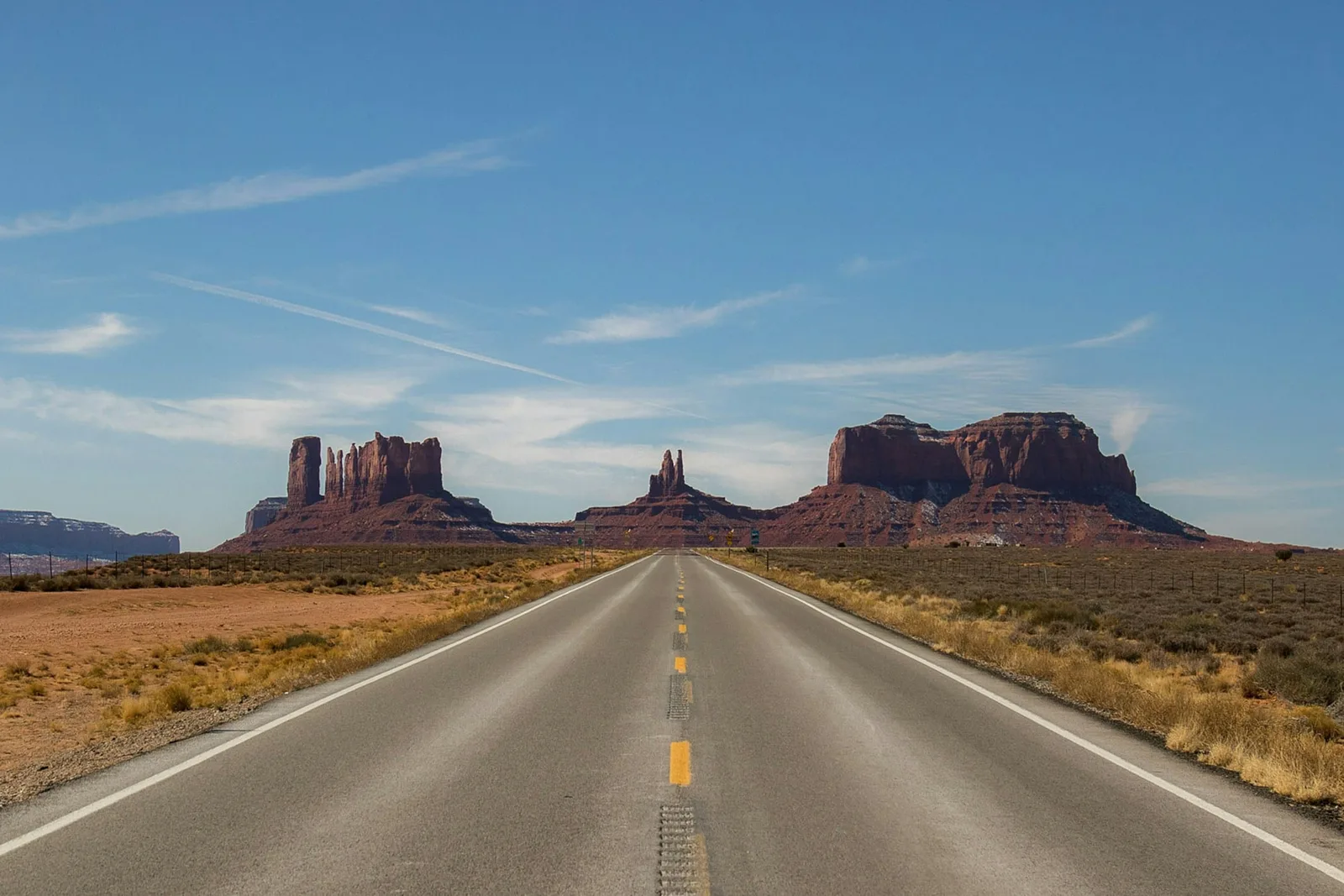 An empty highway running straight toward the sandstone towers of Monument Valley, the desert dry and bright beneath a wide blue sky